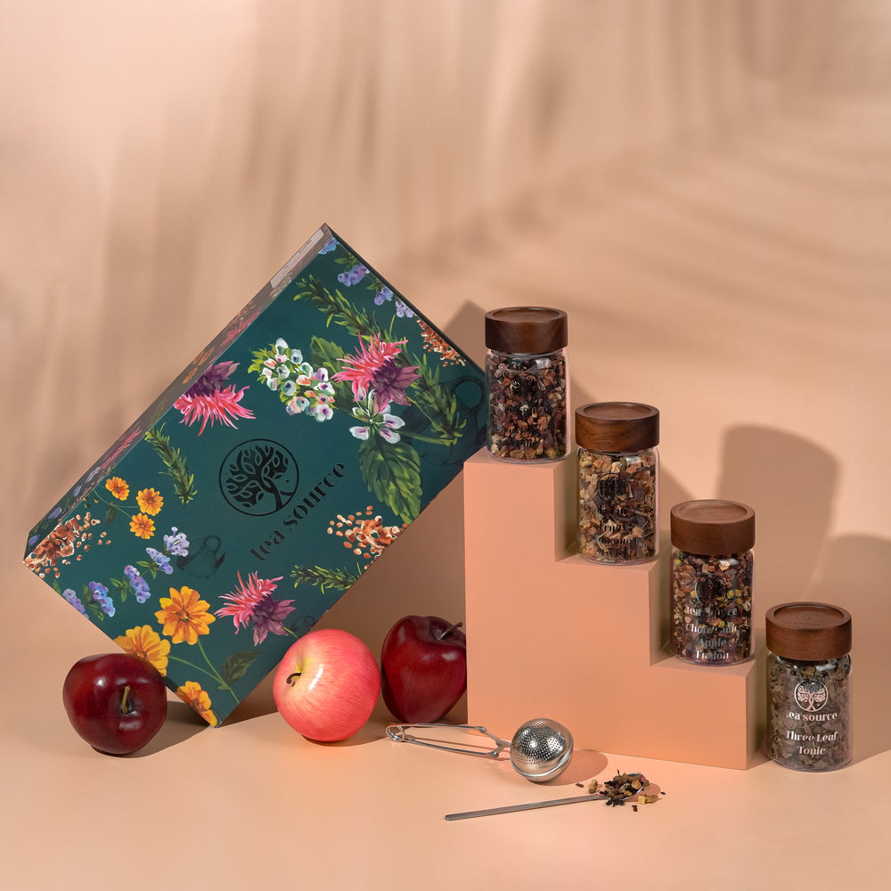 Tea Source Blissful Symphony Tea Gift Set with four glass jars of loose leaf teas (Fruity Cinnamon Twist, Chamomile Apple Fusion, Peach Berry Medley, and Three Leaf Tonic) displayed with apples, a green floral gift box, tea infuser, and spoon.