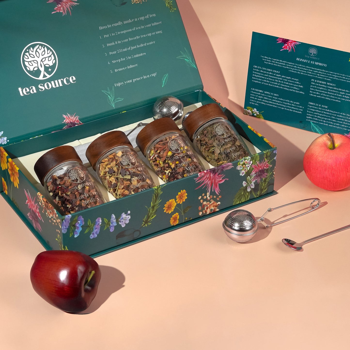 Tea Source Blissful Symphony Tea Gift Set with four glass jars of loose leaf teas (Fruity Cinnamon Twist, Chamomile Apple Fusion, Peach Berry Medley, and Three Leaf Tonic) displayed with apples, a green floral gift box, tea infuser, and spoon.