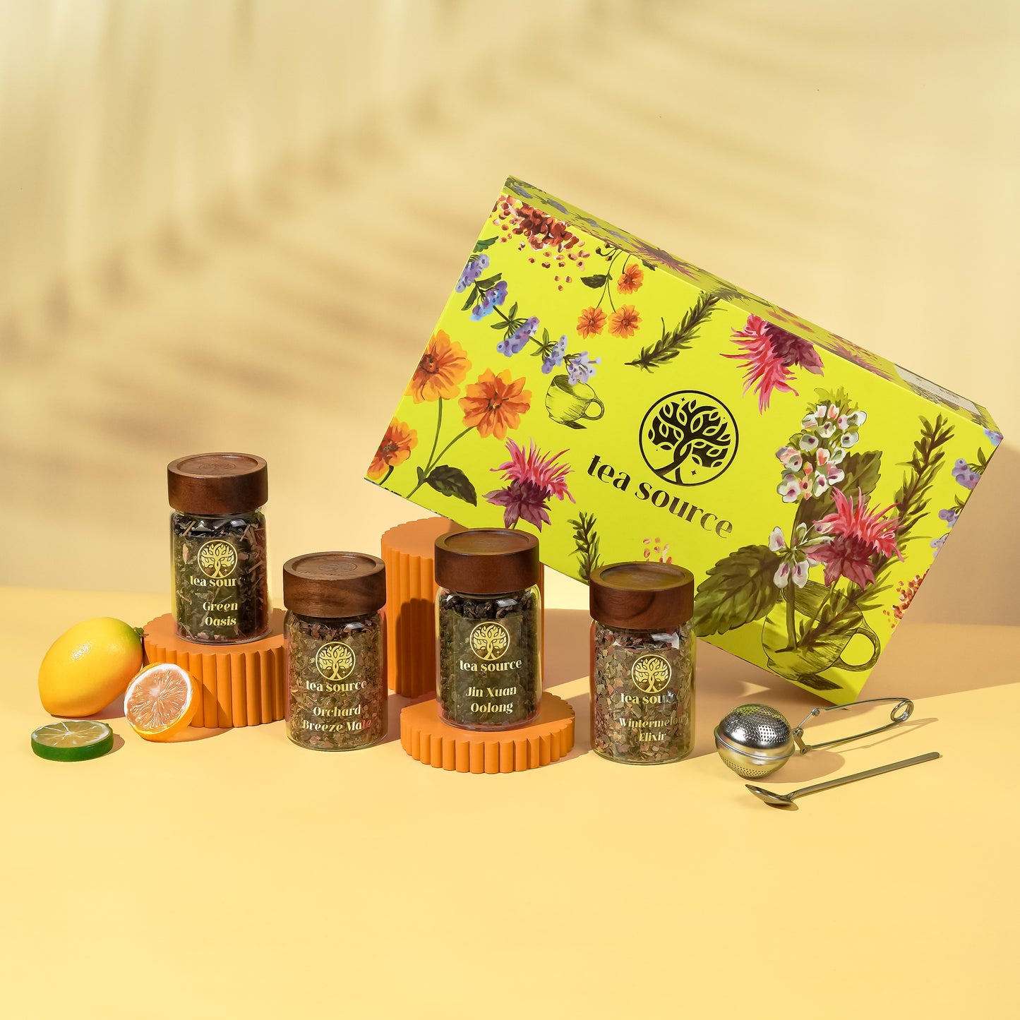 Tea Source Hello New Day Tea Gift Set with four glass jars of loose leaf teas (Green Oasis, Orchard Breeze Maté, Jin Xuan Oolong, and Earl Grey) displayed beside citrus fruits, a floral yellow gift box, and tea infuser tools.
