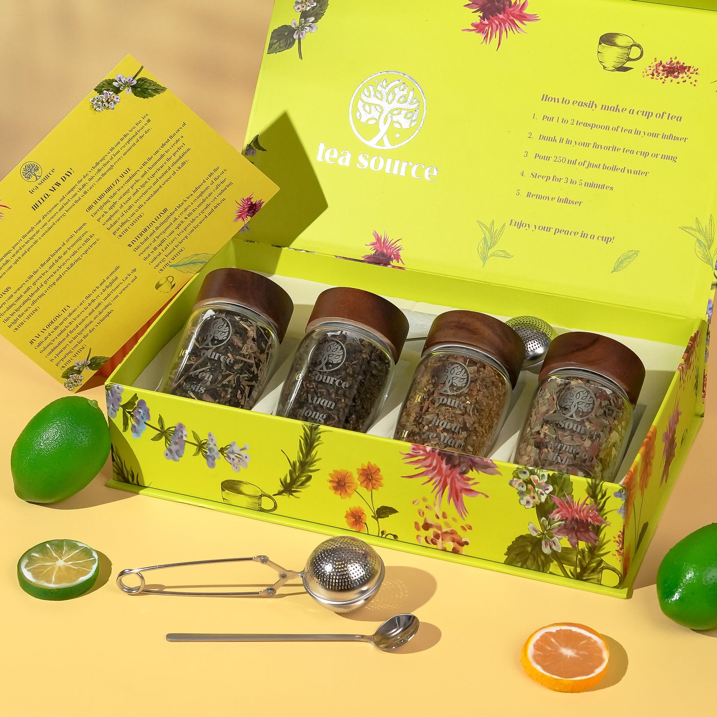 Tea Source Hello New Day Tea Gift Set in an open floral yellow box with four glass jars of loose leaf teas, an instruction card, citrus fruits, and tea infuser tools displayed.