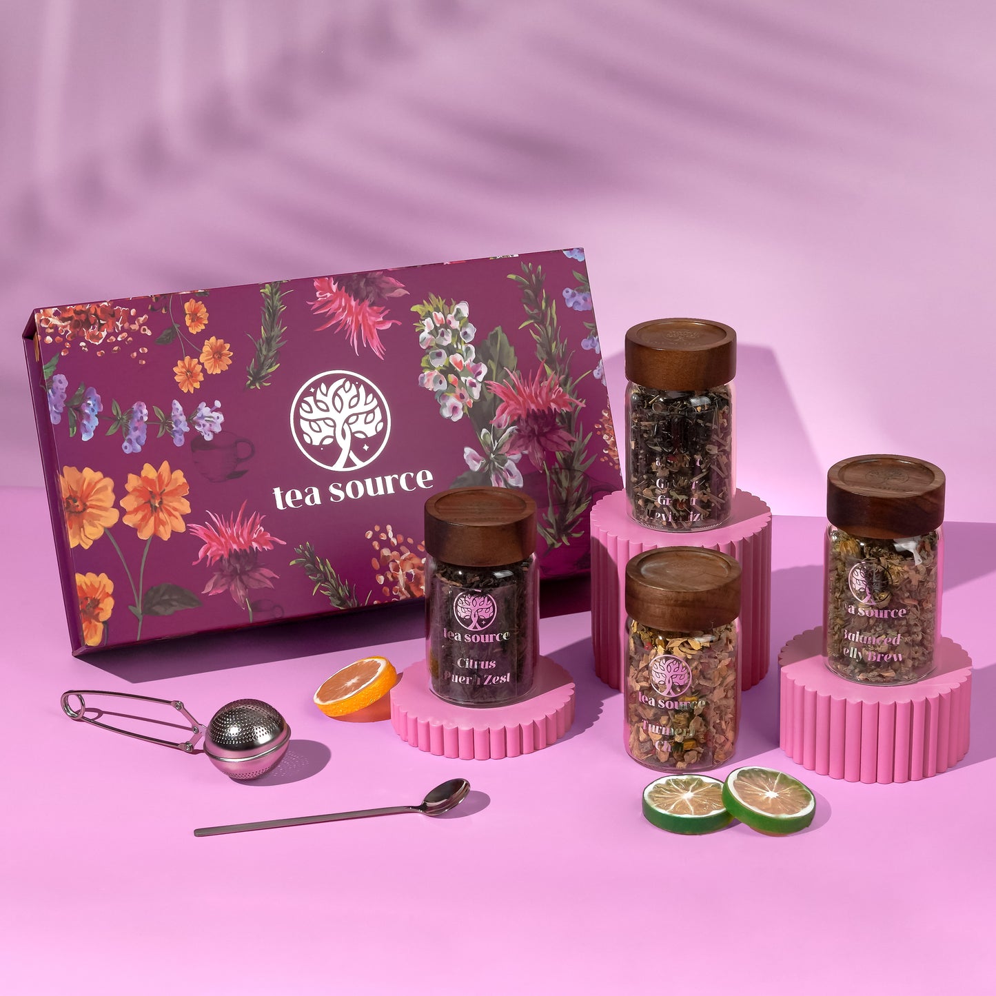 Tea Source Metabolism Boost Tea Gift Set with four glass jars of loose leaf teas (Citrus Puerh Zest, Ginger Green Revitalize, Turmeric Chai, and Balanced Belly Brew) displayed with citrus fruits, a purple floral gift box, and tea infuser tools.