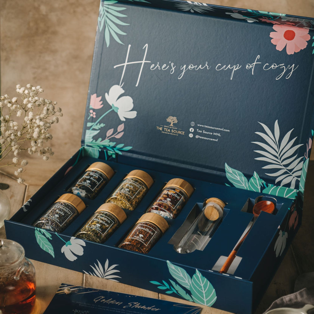 Tea Source Golden Slumber Tea Gift Set in an open blue floral box, featuring six glass jars of loose leaf teas, a glass infuser, wooden spoon, and info card. Designed for relaxation and restful sleep.