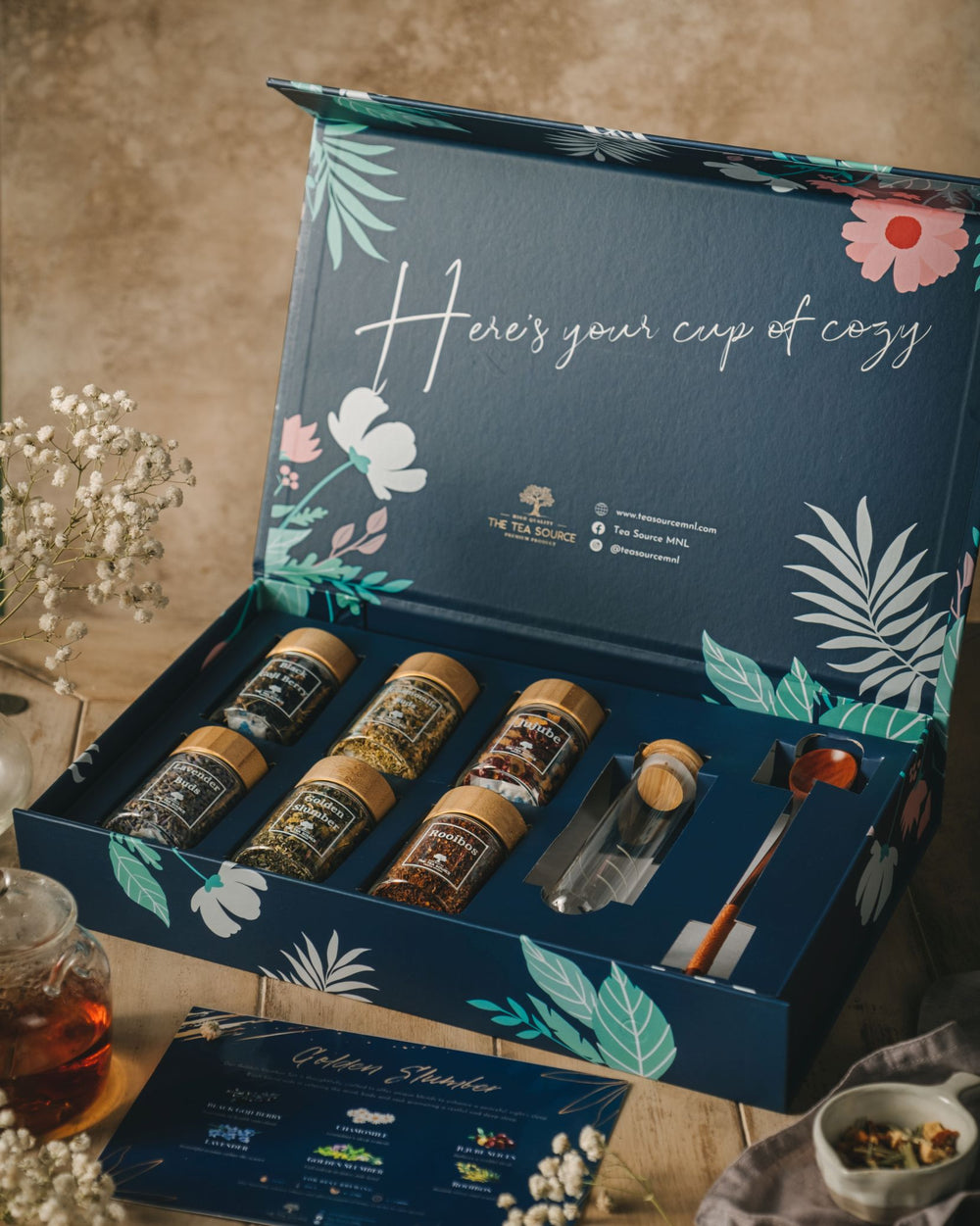 Tea Source Golden Slumber Tea Gift Set in an open blue floral box, featuring six glass jars of loose leaf teas, a glass infuser, wooden spoon, and info card. Designed for relaxation and restful sleep.
