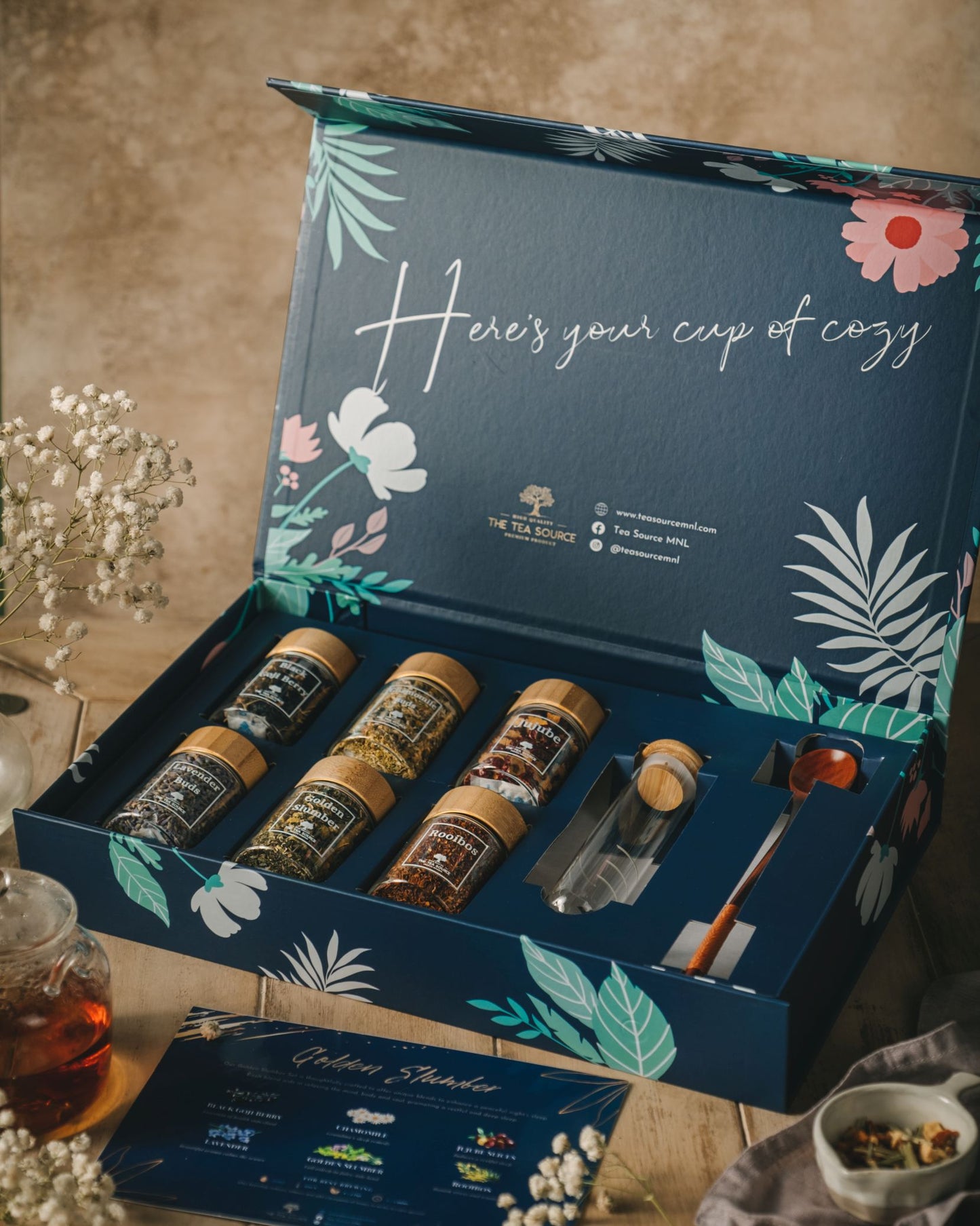 Tea Source Golden Slumber Tea Gift Set in an open blue floral box, featuring six glass jars of loose leaf teas, a glass infuser, wooden spoon, and info card. Designed for relaxation and restful sleep.